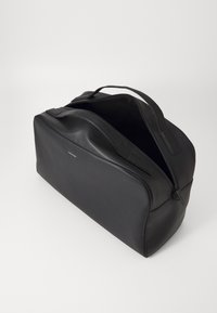 Sac de sport noir en cuir lisse, avec une fermeture éclair et des poignées doubles. Forme rectangulaire avec un intérieur spacieux et un minimum de marquage.