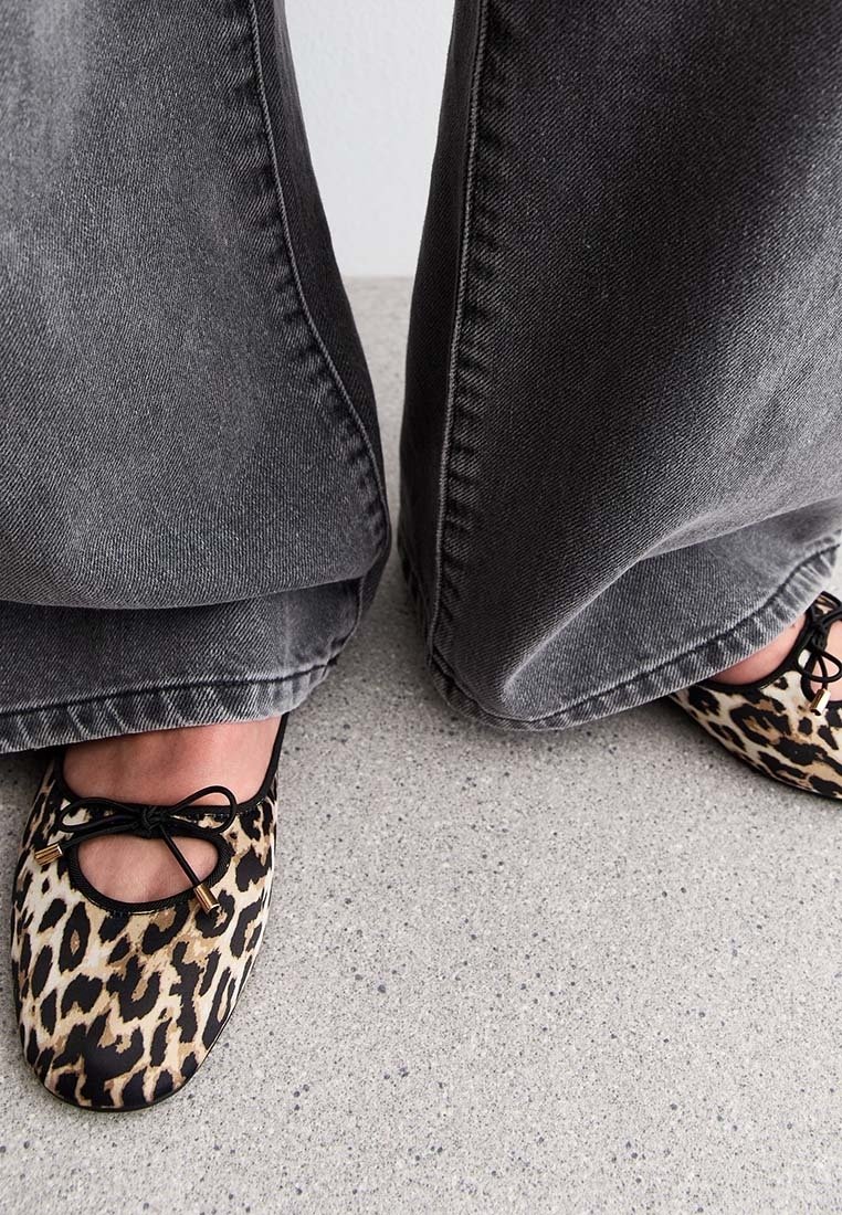 Pieds portant des ballerines imprimé léopard avec des nœuds noirs, associées à un jean gris à jambes larges, debout sur un sol moucheté.