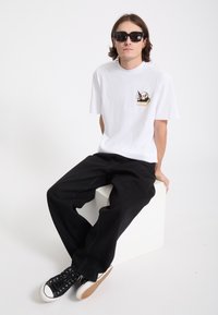 Jeune homme aux cheveux mi-longs portant des lunettes de soleil noires, un t-shirt blanc avec un petit motif de crâne, un pantalon noir et des baskets noires, assis.