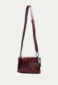 Sac à bandoulière bordeaux en cuir texturé avec fermeture éclair, sangle réglable et accent marron foncé sur le côté.
