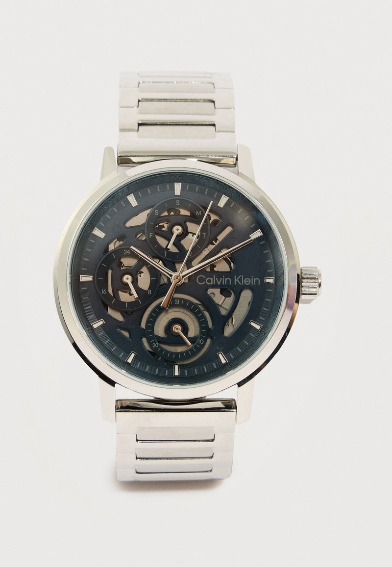 Montre bracelet argentée Calvin Klein avec cadran squelette, trois sous-cadrans et bracelet à maillons métalliques sur fond blanc.
