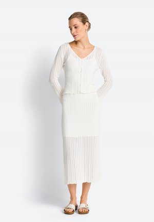 Donna che indossa un cardigan bianco lavorato a maglia con bottoni, gonna abbinata di lunghezza media e sandali bianchi, in piedi davanti a uno sfondo semplice.