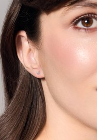 Srebrny kolczyk z małym, przezroczystym kamieniem szlachetnym w kwiatowym wzorze. Kolczyk jest prosty i elegancki, osadzony na gładkiej skórze.