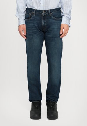Jeans en denim bleu foncé avec une coupe droite, cinq poches et des détails de coutures subtils, associés à des baskets noires.