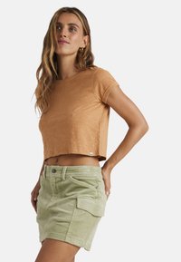 T-shirt corta marrone realizzata in tessuto morbido, abbinata a pantaloni shorts in velluto a coste verde chiaro con tasche frontali e chiusura con bottoni.