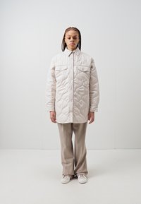 Jachetă căptușită, matlasată, în bej deschis, cu închidere cu nasturi, două buzunare pe piept și o croială lejeră. Asortată cu pantaloni bej și încălțăminte albă.