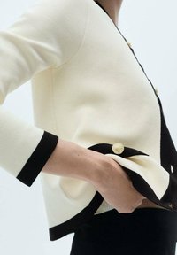 Osoba oblečená v krémovém a černém kardigánu se zlatými knoflíky, s rukou zasunutou v kapse kardigánu, ukazující texturovanou pletenou látku.