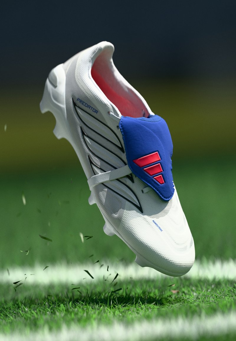 Witte voetbalschoenen met blauwe accenten, een textuurovervlak, zwarte strepen en rode details. Zwevend boven gras waarbij de grassprieten worden verstoord.