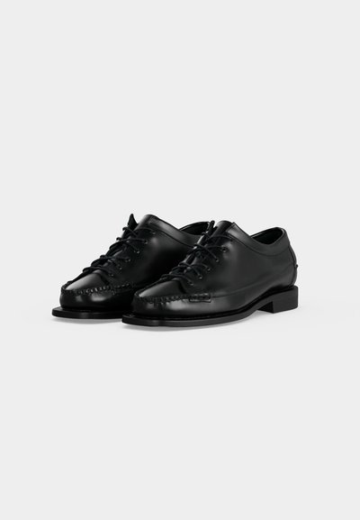 Paire de chaussures habillées décontractées en cuir noir à lacets, avec couture moc toe et talons bas, sur un fond clair uni.