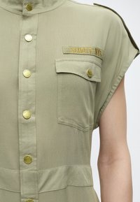 Αμάνικο χακί πουκάμισο με χρυσά κουμπιά, τσέπη με καπάκι στο στήθος και κεντημένη ετικέτα "SCHOTT-NYC" πάνω από την τσέπη στην αριστερή πλευρά.