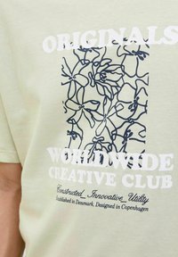 Lys grøn t-shirt med rund hals, prydet med et marineblåt blomstermotiv og hvid tekst, der siger "ORIGINALS WORLDWIDE CREATIVE CLUB."