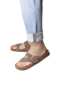 Brown suede sandals with crossover straps and a buckle. Cork sole and rubber outsole. Worn with rolled-up light denim jeans.
