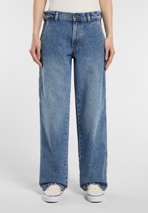 A person wears loose-fitting blue jeans, styled with a plain top and light shoes, standing against a neutral background.