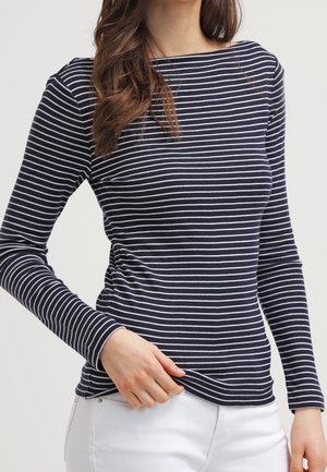 Woman wearing a navy blue long-sleeve shirt with white horizontal stripes, paired with white pants, standing against a plain background.