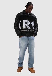 Czarna kurtka z polaru i skóry z białym graficznym napisem; jasnoniebieskie dżinsy; brązowe buty robocze; luźny krój, kontrastująca faktura materiału.