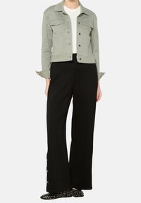 Kobieta w jasnozielonej kurtce zapinanej na guziki, białej bluzce, czarnych szerokich spodniach z bocznymi guzikami oraz czarnych płaskich butach z ćwiekami.