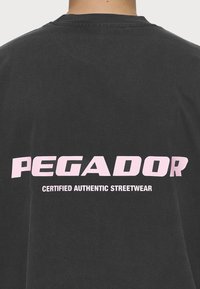 Fekete pamut póló, nagy rózsaszín felirattal a hátsó részén, ami a következőt olvasható: "PEGADOR" és alatta "CERTIFIED AUTHENTIC STREETWEAR".