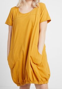 Senapsgul oversized klänning med korta ärmar, rundad halsringning och stora sidofickor; har en rynkad fåll och mjukt, texturerat tyg.