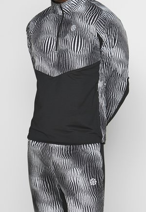 Mann trägt schwarzen und weißen Langarmpullover und passende Hose mit abstraktem vertikal gestreiftem Muster und Reißverschluss am Kragen.