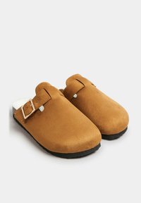 Zuecos de ante marrón con una forma curva, hebilla ajustable, forro de piel sintética blanca y suela de goma negra. Sin patrones ni acentos adicionales.