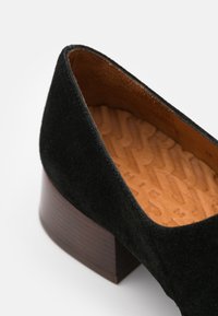 Chaussure en suede noire avec un talon en bois de hauteur moyenne. L'intérieur présente une doublure en tan texturée. Bout arrondi et finition extérieure lisse.