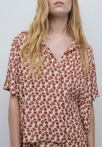 Kortærmet skjorte i creme med et rødt blomstermønster, der har knaplukning foran og en afslappet pasform. Blødt stof med en let tekstur.