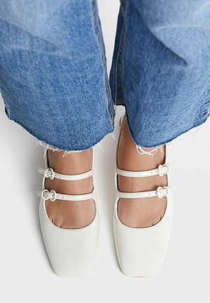 Des pieds portant des ballerines blanches à double bride avec un bout carré, associées à un jean en denim bleu à ourlet effiloché sur un fond blanc.
