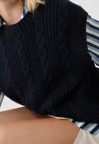 Närbild av en person som bär en marinblå kabelstickad ärmlös tröja över en blå och vit randig långärmad skjorta samt beige shorts.