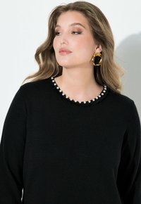 Schwarzer Pullover mit rundem Ausschnitt, verziert mit perlenähnlichen Akzenten und einer strukturierten Fransenbordüre. Das Modell trägt große goldene Creolen.