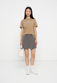 Μπλούζα t-shirt σε ταμπά χρώμα με εκτύπωση, γκρι πλισέ φούστα με κουμπιά, λευκά αθλητικά παπούτσια και μια μικρή λευκή τσάντα. Το μοντέλο στέκεται σε απλό φόντο.