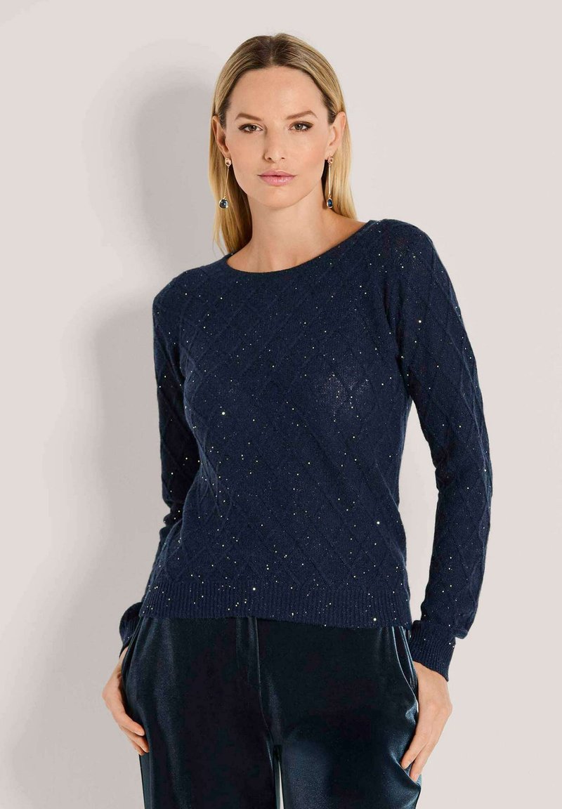 Frau, die einen dunkelblauen Strickpullover mit subtilen Glitzerakzenten und passenden dunkelblauen Hosen trägt, vor einem schlichten hellen Hintergrund steht.
