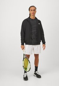 Zwarte sportjack over een gestreept donker shirt, witte shorts en zwarte tennisschoenen. Houdt een tennisracket vast met een geel en zwart ontwerp.