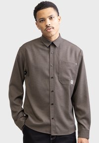 Brown checkered long-sleeve button-up shirt, featuring a pointed collar, a chest pocket, and black buttons. Made of textured fabric.