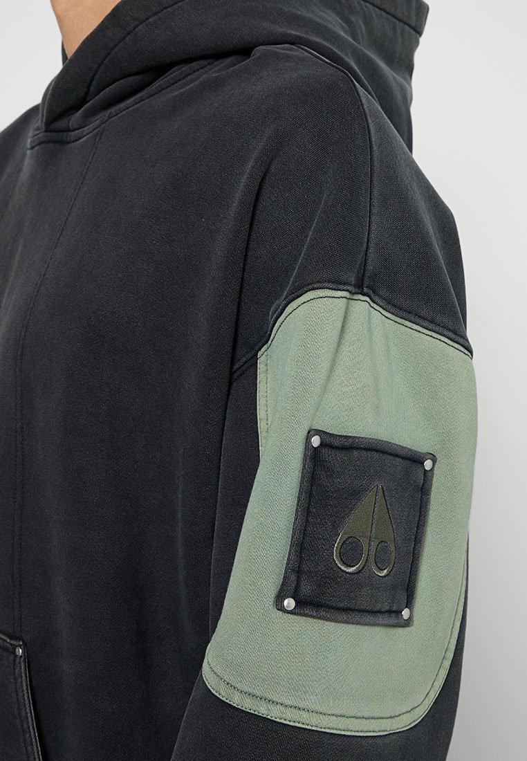 Schwarzer Hoodie mit einer strukturierten Oberfläche, ausgestattet mit einem olivgrünen Ärmelpatch, der mit silbernen Nieten befestigt ist, und einem Scheren-Graphic. Aus weichem Baumwollmaterial.