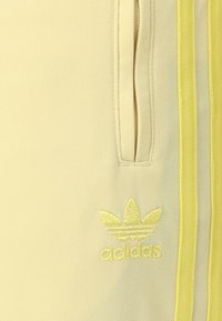 Világ sárga sportnadrág, két oldalsó zsebbel, kontrasztos sárga csíkokkal és hímzett Adidas logóval ugyanabban a színben.
