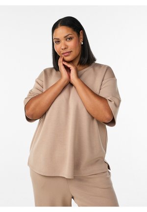 Moteris su vidutinio tamsumo oda ir tiesiais juodais plaukais, vilkinti smėlio spalvos laisvalaikio viršutinį drabužį ir kelnes, rankos švelniai liečia veidą, neutralus fonas.