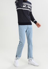 Marineblauwe gebreide trui met witte wintermotieven, gedragen met lichtblauwe jeans en witte sneakers. De trui heeft ribgebreide manchetten en zoom.