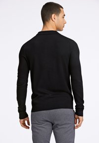 Svart strikket genser med krage, tettsittende silhuett, og ribbestrikkede mansjetter og nederkant, sammen med grå joggebukser. Bakview.