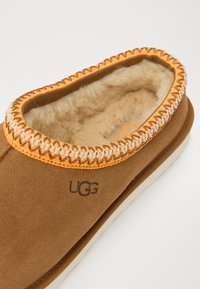 Rjavi semišna copatka z notranjim krznom, oranžni šivi in logotipom UGG vtisnjenim na strani. Okrogla oblika s oblazinjenim podplatom.