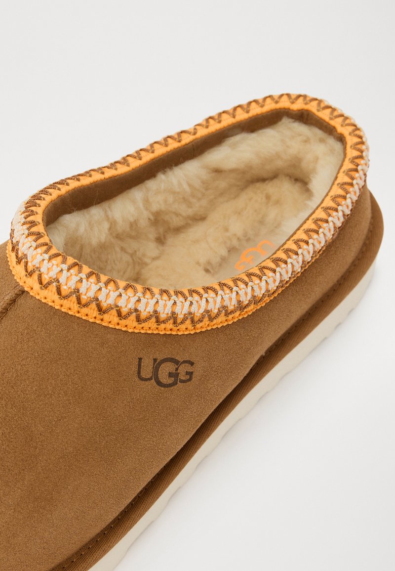 Pantofole in suede marrone con rivestimento in pelliccia, dettagli arancioni a punto e logo UGG impresso sul lato. Forma rotonda con suola imbottita.