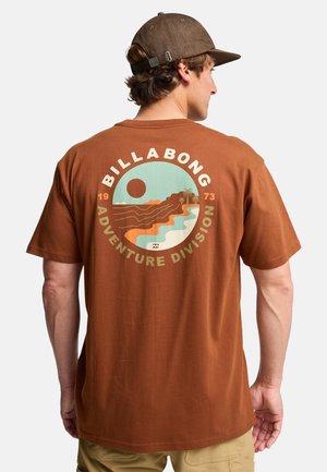 Bruine katoenen t-shirt met een cirkelvormige grafiek in blauwgroen en oranje met een ontwerp van de zon, een berg en een golf. De tekst luidt "BILLABONG ADVENTURE DIVISION."