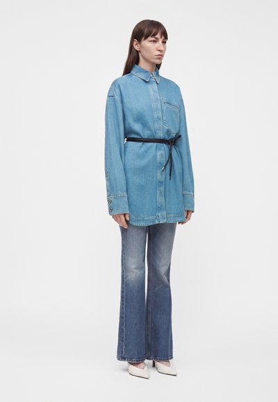 Femme aux longs cheveux bruns portant une chemise en jean bleu clair ample ceinturée à la taille, un jean bleu délavé et des chaussures blanches pointues.