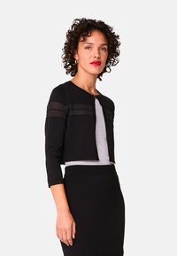 Zwarte cropped cardigan met driekwart mouwen, voorzien van doorzichtige panelen. Gecombineerd met een grijze getailleerde top en een zwarte kokerrok.