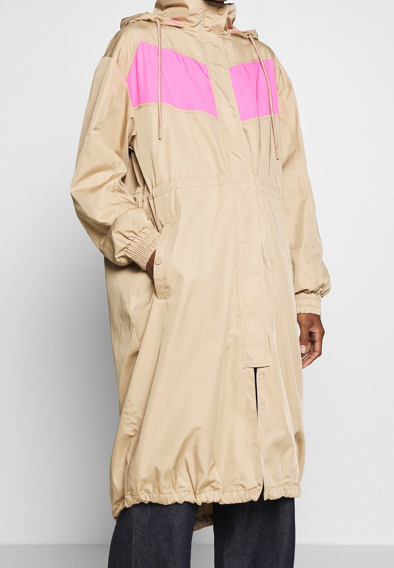 Vaaleanbeige hupparitakki, jossa on vaaleanpunaiset rintapaneelit, joustavat hihansuut, etutaskut ja säädettävät nyörit, käytetty tummien housujen päällä.