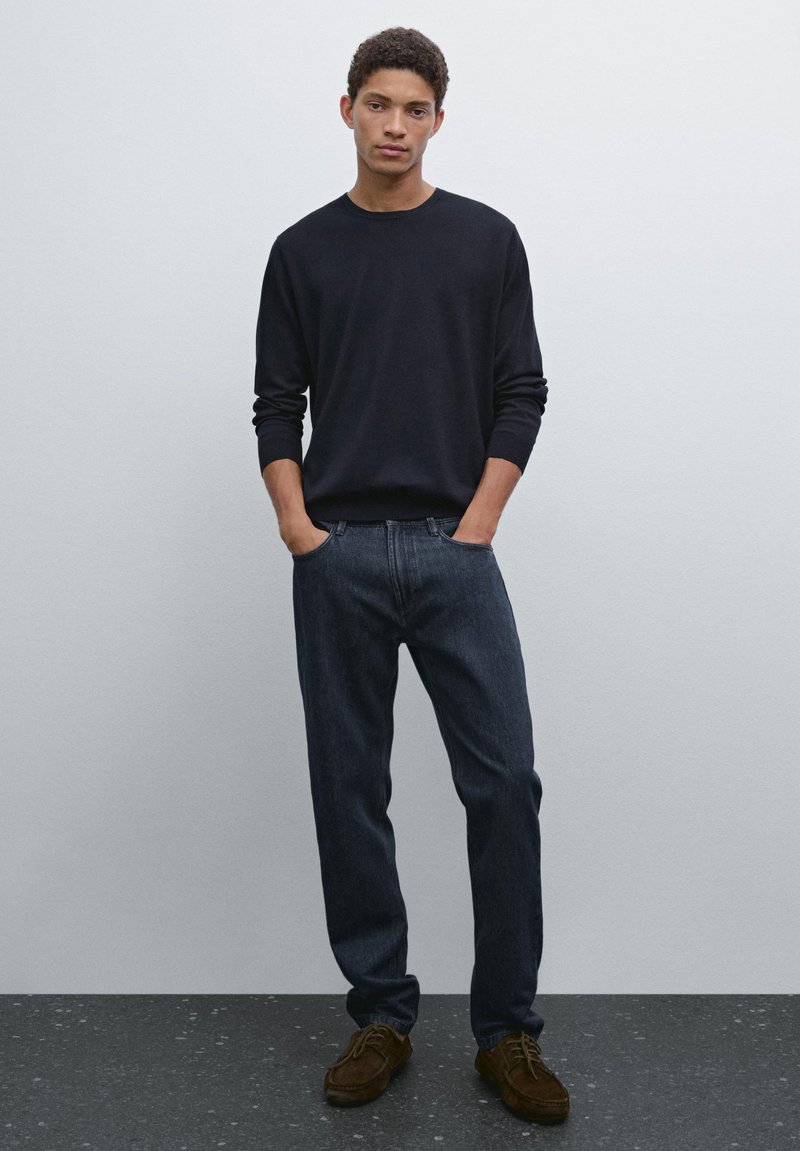 Dark blue knit sweater, relaxed fit dark jeans, and brown lace-up shoes. Smooth fabric with ribbed cuffs, simple design. Plain background.
