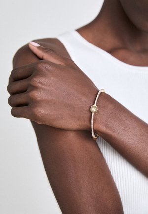 Gouden armband met een textiele band, voorzien van een cirkelvormige schijfvertoon met een logo en een klein kraaldetail.