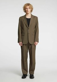 Brązowy garnitur na miarę z jednorzędową marynarką i spodniami o prostych nogawkach, wykonany z gładkiej tkaniny, noszony na czarnej bluzce, uzupełniony czarnymi butami.