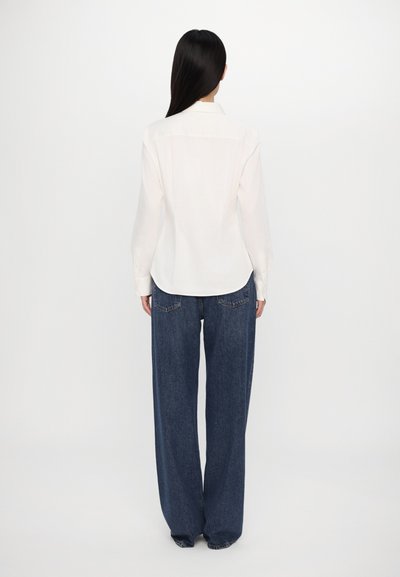 White button-up shirt with long sleeves, paired with loose-fitting dark blue jeans, displayed from the back against a plain background.