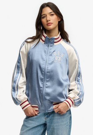 Młoda kobieta nosząca błękitno-kremową satynową kurtkę bomberkę z mankietami i kołnierzem w czerwone paski, wyszywaną napisem "U.S.A 83 Superdry."