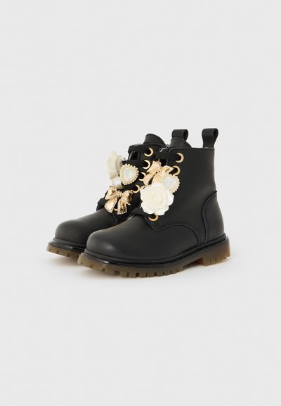 Botas de tobillo de cuero negro con ojetes dorados, cordones negros y encantos decorativos en forma de flor blanca, corazón y lazo en los cordones.